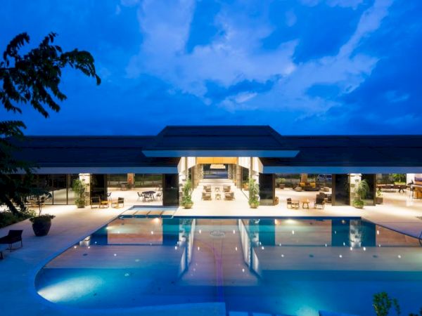 A modern building with a large, illuminated swimming pool and outdoor seating area at dusk, surrounded by trees and featuring an open, inviting design.