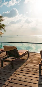 Poolside deck with lounge chairs and umbrellas, overlooking a serene ocean and tropical landscape under a partly cloudy sky.