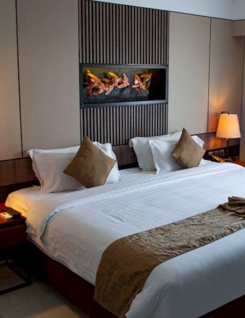 A well-lit hotel room with a large bed, two bedside tables with lamps, pillows, and a wall-mounted fireplace.