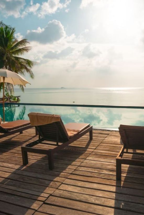 The image shows a serene poolside with lounge chairs, umbrellas, and palm trees. It overlooks a calm sea under a cloudy sky during daylight.