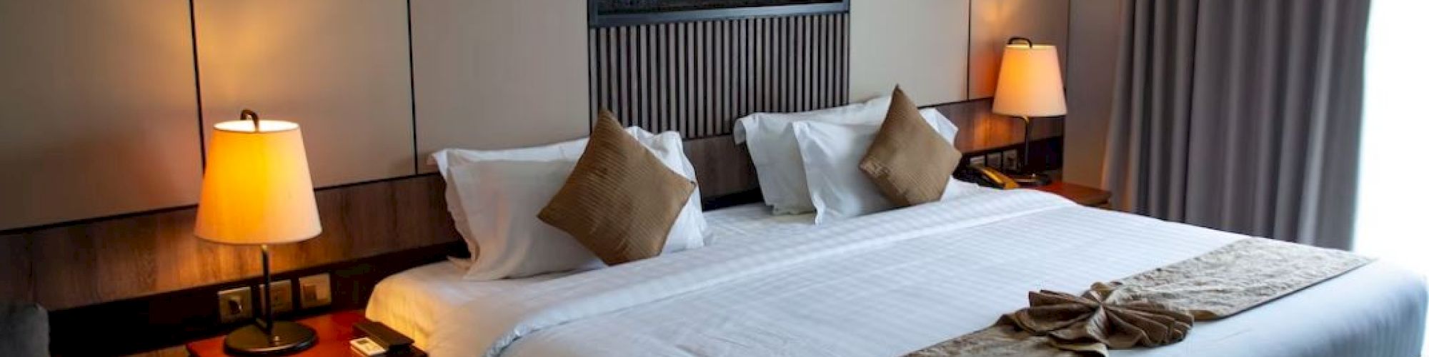 A cozy hotel room with a large, neatly made bed, bedside lamps, and warm lighting. The room has modern decor and a window with curtains.