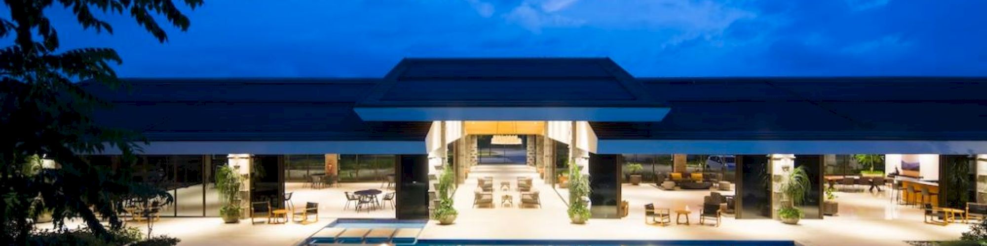A luxurious building with an open layout and a large illuminated pool in front, set against a twilight sky.