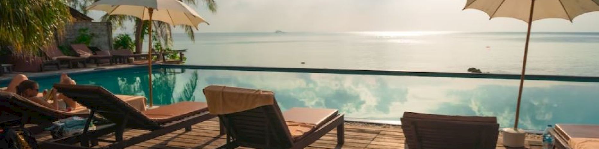 A serene poolside view with loungers and umbrellas, overlooking a tranquil body of water under a partly cloudy sky, framed by palm trees.