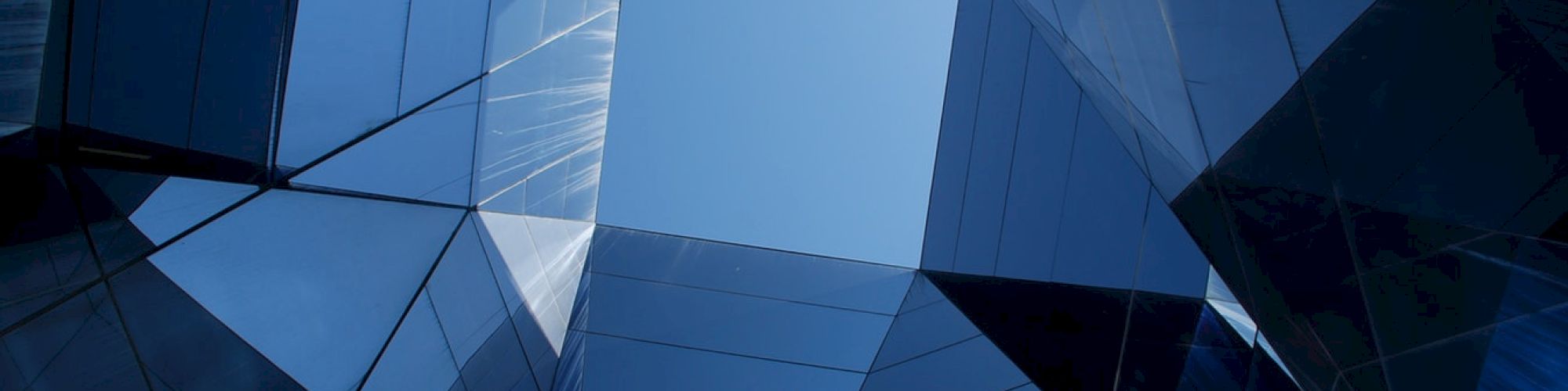The image shows an upward view of a modern building with a geometric design made of reflective glass and metal, revealing a clear section of sky.