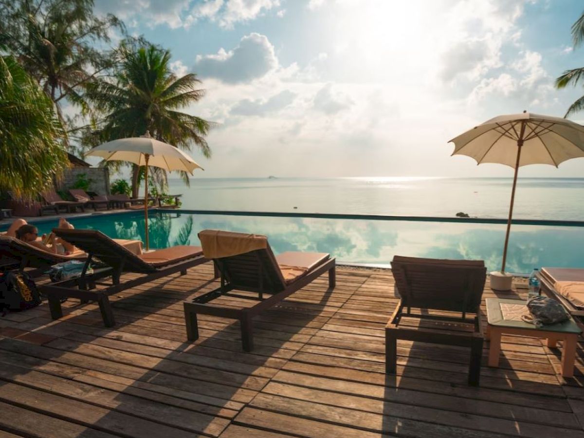 A serene poolside scene with lounge chairs, umbrellas, and a view of the ocean under a partly cloudy sky, framed by palm trees.