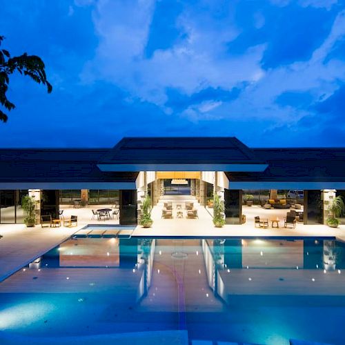 A luxurious pool area with a modern open building in the background set against a twilight sky, illuminated by ambient lighting.