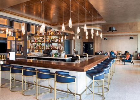 The image shows a modern bar with a wooden counter, ample seating, a fully stocked back bar, pendant lights, and a sitting area in the background.