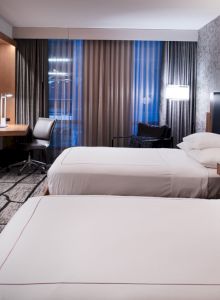 This image shows a modern hotel room with two neatly made beds, a flat-screen TV, a desk, and large windows with curtains.