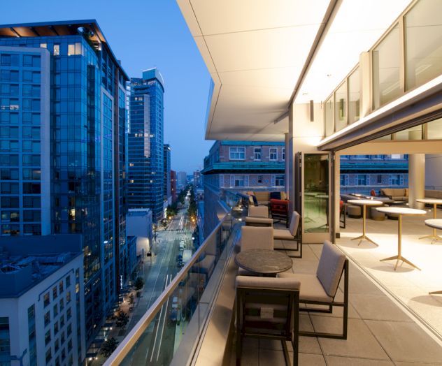 A modern balcony view of a cityscape at dusk, with buildings, streetlights, and a sleek outdoor seating area with tables and chairs.