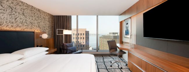 A modern hotel room with a large bed, wall-mounted TV, desk, chair, and a city view through floor-to-ceiling windows is shown.