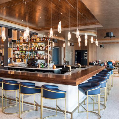 This image shows a modern bar with blue cushioned stools, a well-stocked bar, hanging pendant lights, and a casual seating area in the background.
