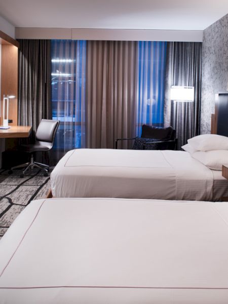 The image shows a modern hotel room with two neatly made beds, a flat-screen TV, a desk with a chair, and large windows with blinds.