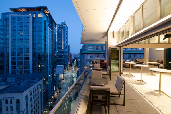 The image shows a modern cityscape at twilight, with high-rise buildings and a stylish outdoor patio space featuring chairs, tables, and a glass railing.