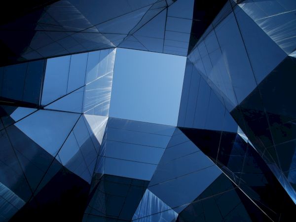 The image shows a view looking up at modern, reflective glass building structures with a geometric design, capturing the blue sky in the center.