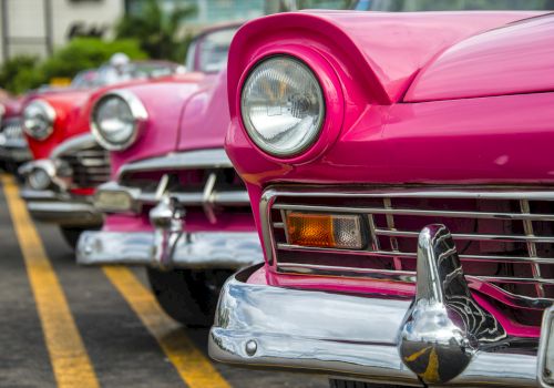 The image shows a row of colorful vintage cars parked in a lot, with the focus on a pink one in the front and the others slightly blurry.