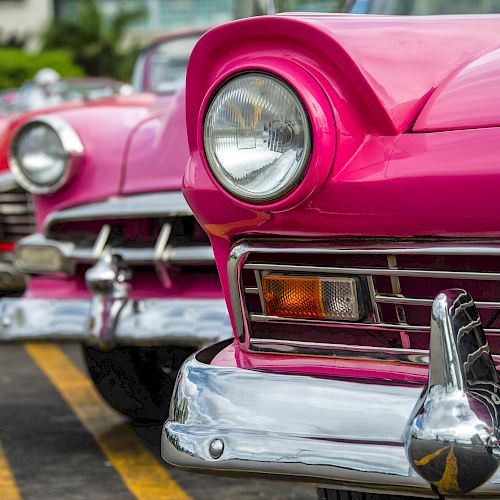 The image shows a row of colorful vintage cars parked in a lot, with the focus on a pink one in the front and the others slightly blurry.