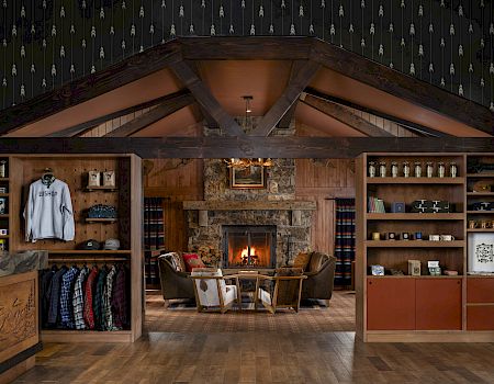 A cozy wooden lodge interior with a lit stone fireplace, couches, rugs, and shelves of gear and clothes.