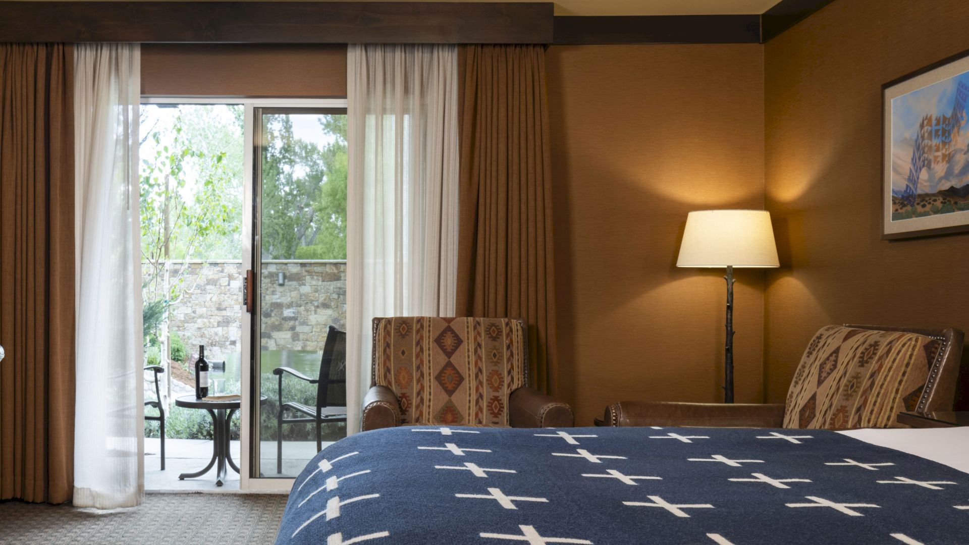 A cozy hotel room with a bed, nightstands, lamps, armchairs, a desk, and a sliding glass door leading to a small patio with outdoor furniture.