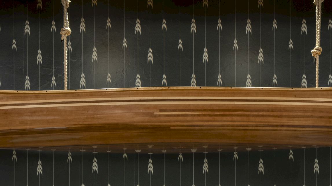 A wooden canoe is suspended from ropes against a patterned black wall featuring white arrow designs.