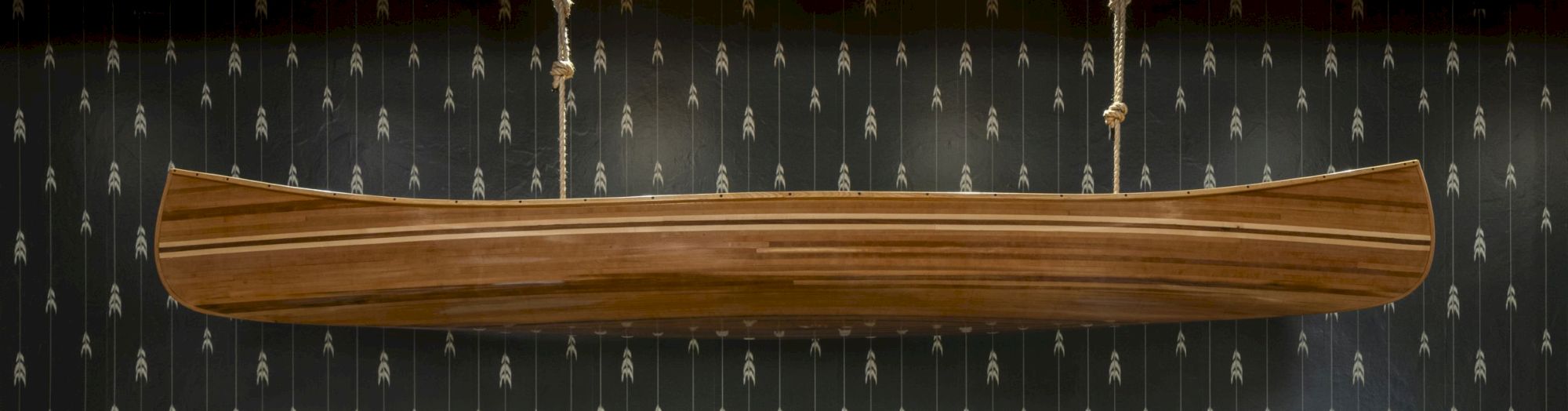 A wooden canoe is suspended from ropes against a patterned black wall featuring white arrow designs.