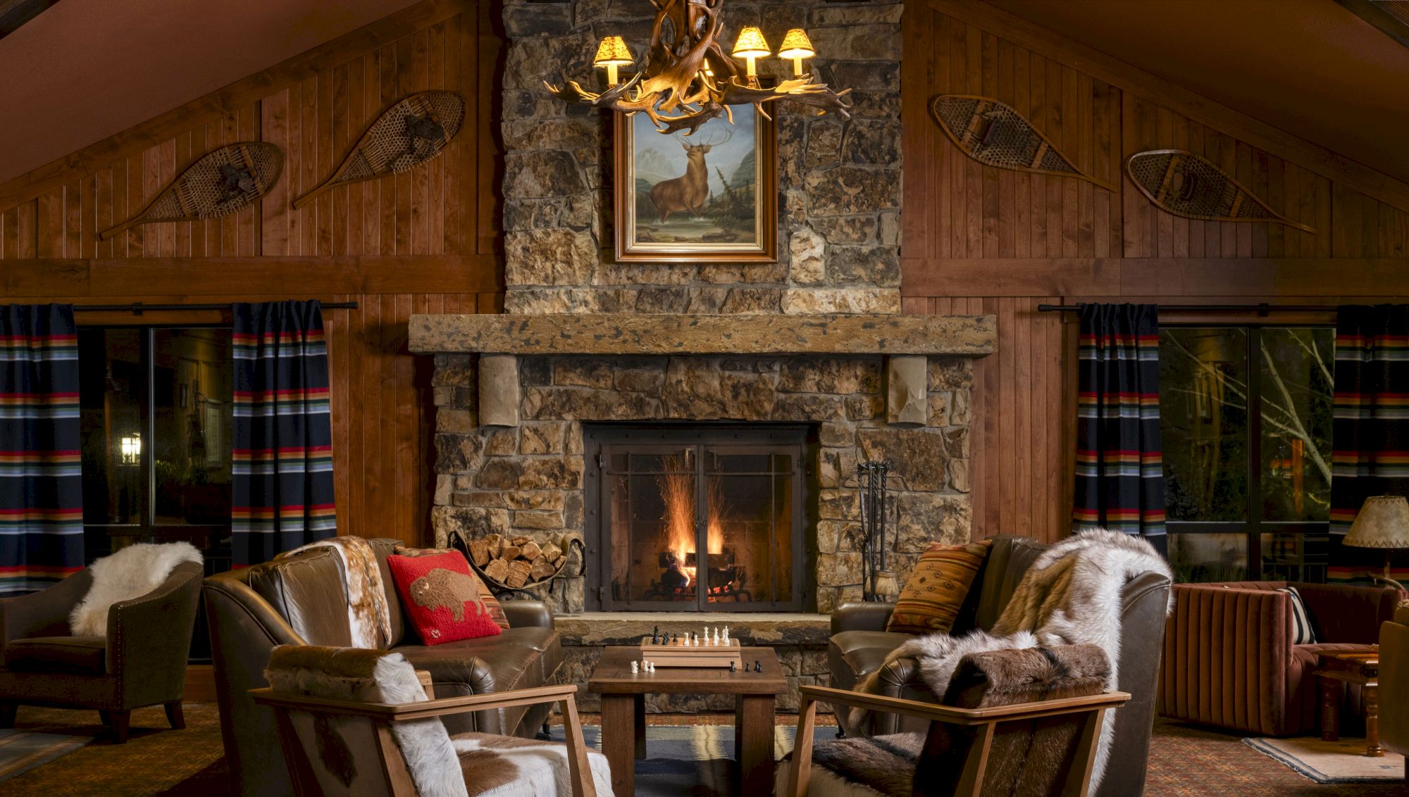 A cozy, rustic living room with a stone fireplace, wooden walls, antler chandelier, and comfortable seating arranged around a chess table.