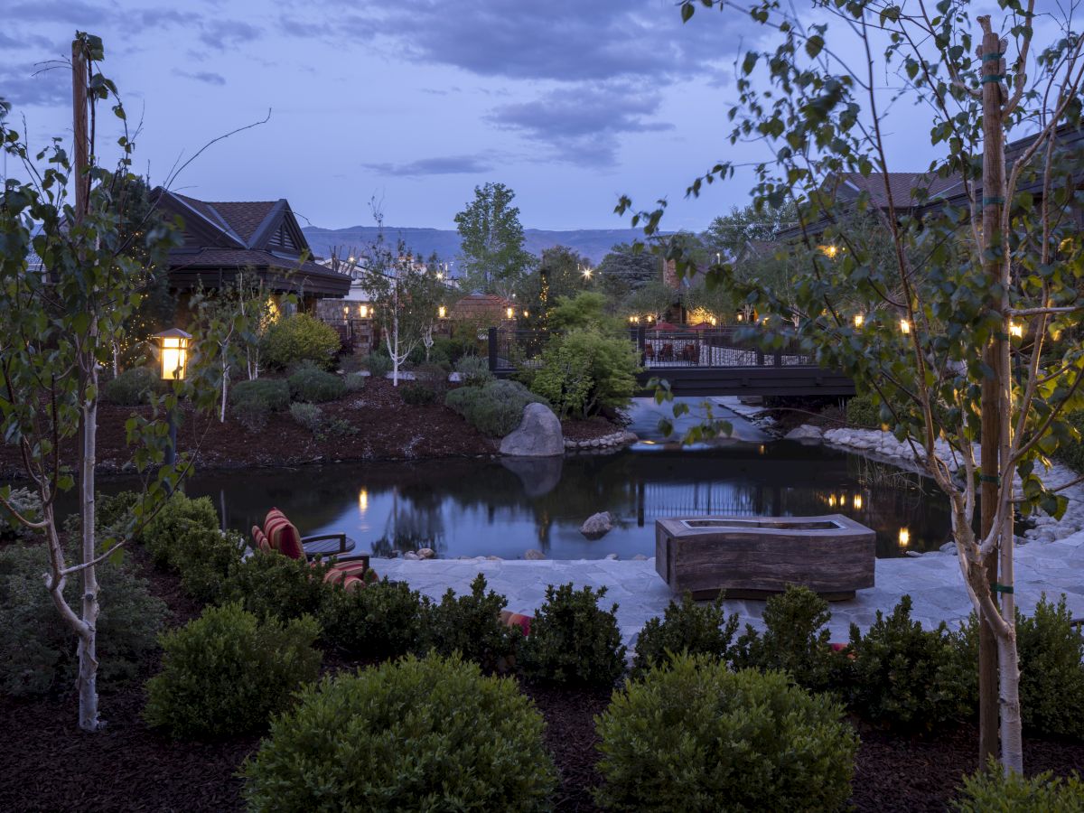 The image shows a serene garden at dusk with a pond, trees, and lit pathways, creating a peaceful atmosphere.