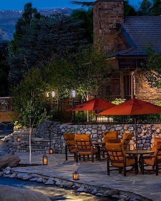 An outdoor patio with tables, chairs, and red umbrellas by a lit pathway and a pond near a stone building with scenic nature views in the evening.