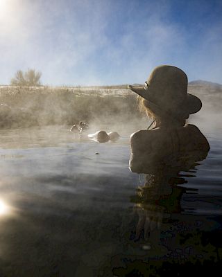 A person wearing a hat is bathing in a steamy outdoor hot spring under a bright sky, with another person visible in the background.