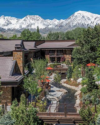 A lodge with a stone exterior is surrounded by lush greenery, with snow-capped mountains in the background and a small stream running through the courtyard.