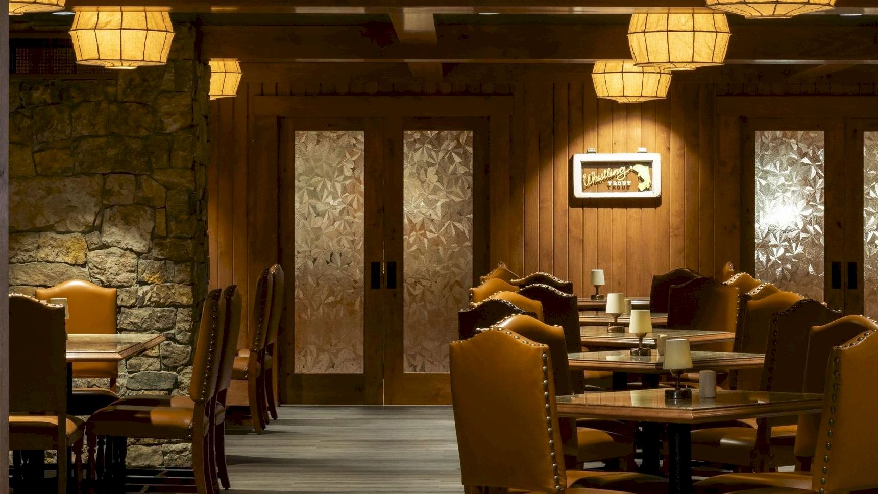 An elegant, dimly lit restaurant interior with wooden decor, cushioned chairs, lantern-style lights, and frosted glass doors at the back.