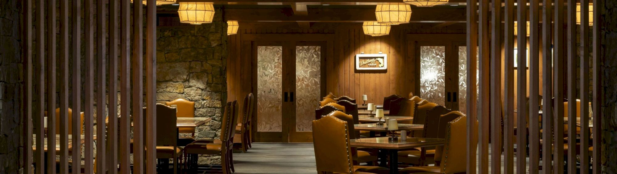 An elegant, dimly lit restaurant interior with wooden decor, cushioned chairs, lantern-style lights, and frosted glass doors at the back.