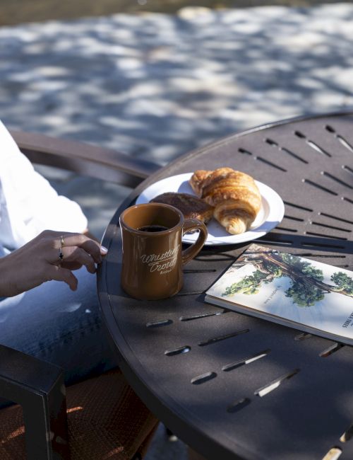 A person is seated at an outdoor table with a coffee mug, a plate of croissants, and what appears to be a book or magazine on the table.