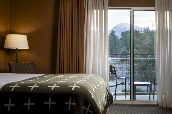 A cozy hotel room with a bed, bedside lamp, and curtains, opens to a balcony with chairs, offering a scenic view of mountains.