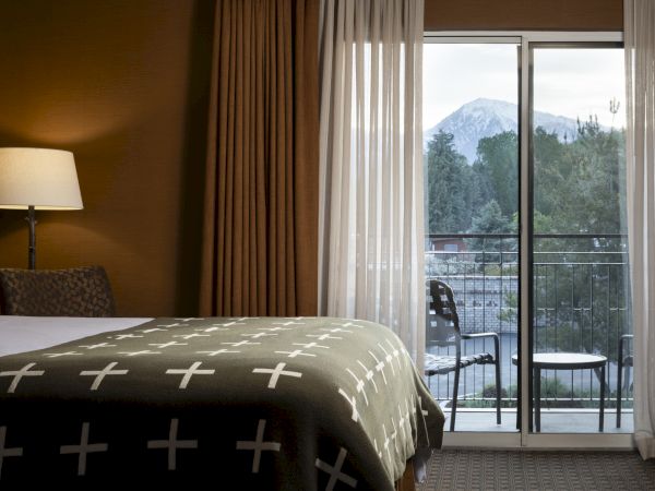 A cozy hotel room with a bed, bedside lamp, and curtains, opens to a balcony with chairs, offering a scenic view of mountains.