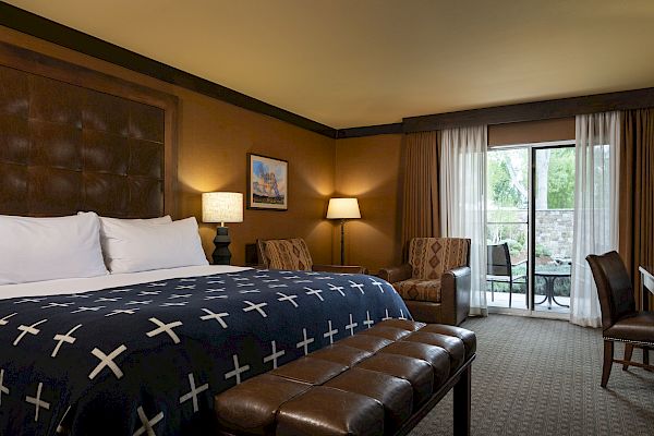 The image shows a cozy hotel room with a large bed, a padded bench, armchairs, a desk, lamps, a painting, and a patio door with curtains leading outside.