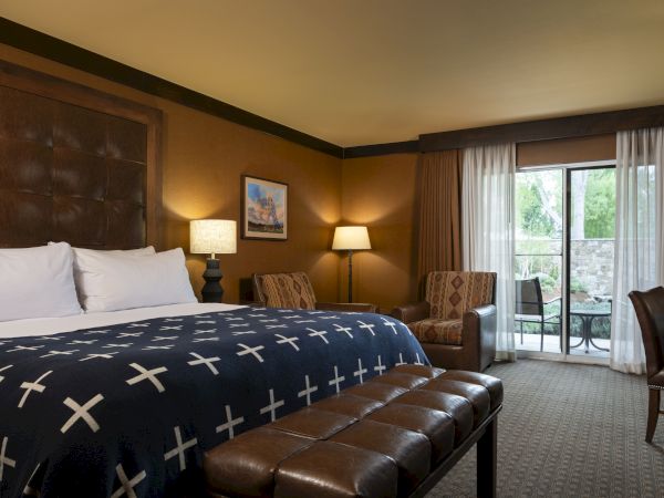 The image shows a cozy hotel room with a large bed, a padded bench, armchairs, a desk, lamps, a painting, and a patio door with curtains leading outside.