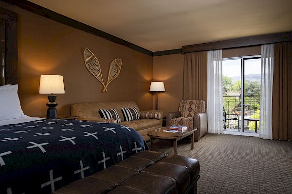 A cozy hotel room features a bed, sofa, armchair, lamps, and a glass door leading to a balcony with a scenic view.