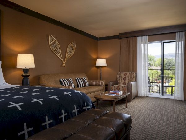 A cozy hotel room features a bed, sofa, armchair, lamps, and a glass door leading to a balcony with a scenic view.