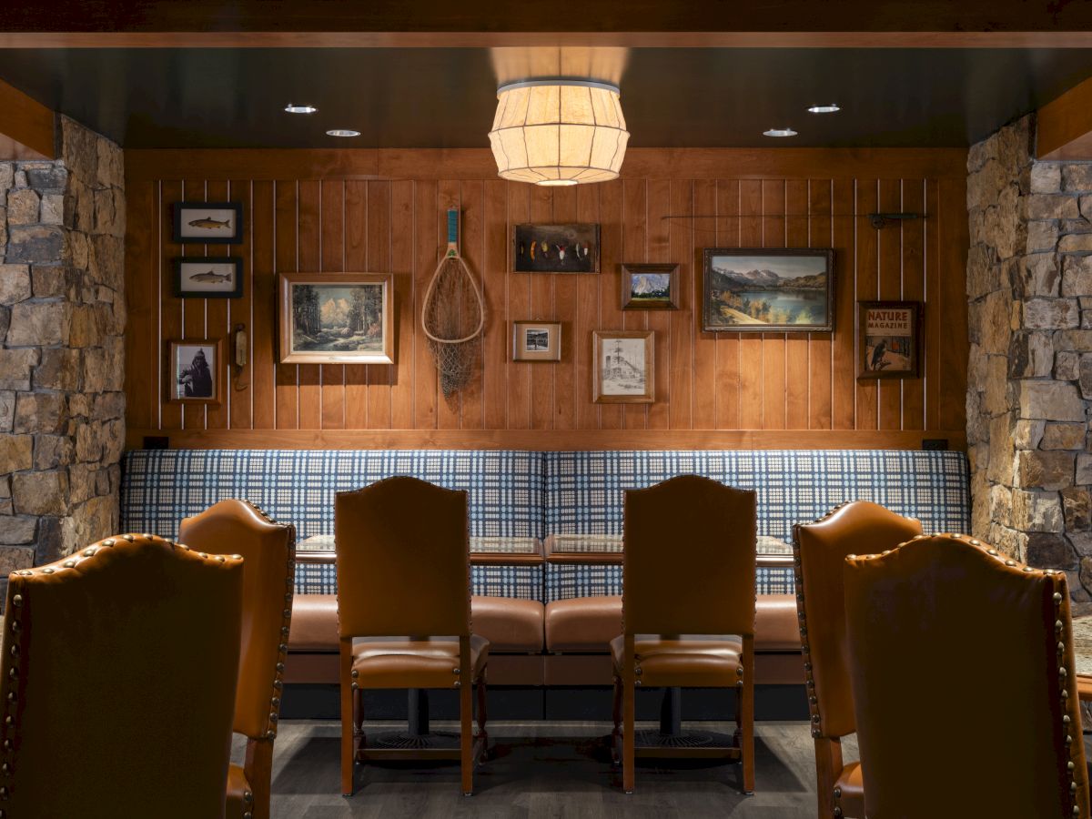 A cozy dining area with stone walls, wooden paneling, framed pictures, and six chairs around a table, illuminated by a hanging lamp.