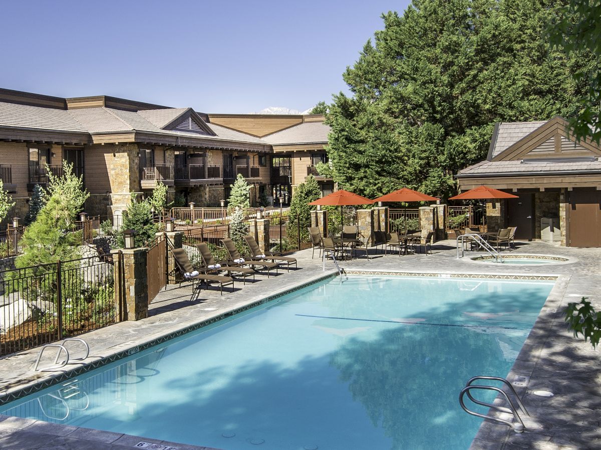 An outdoor hotel courtyard with a turquoise swimming pool, lounge chairs, red umbrellas, and a brick building surrounded by trees.