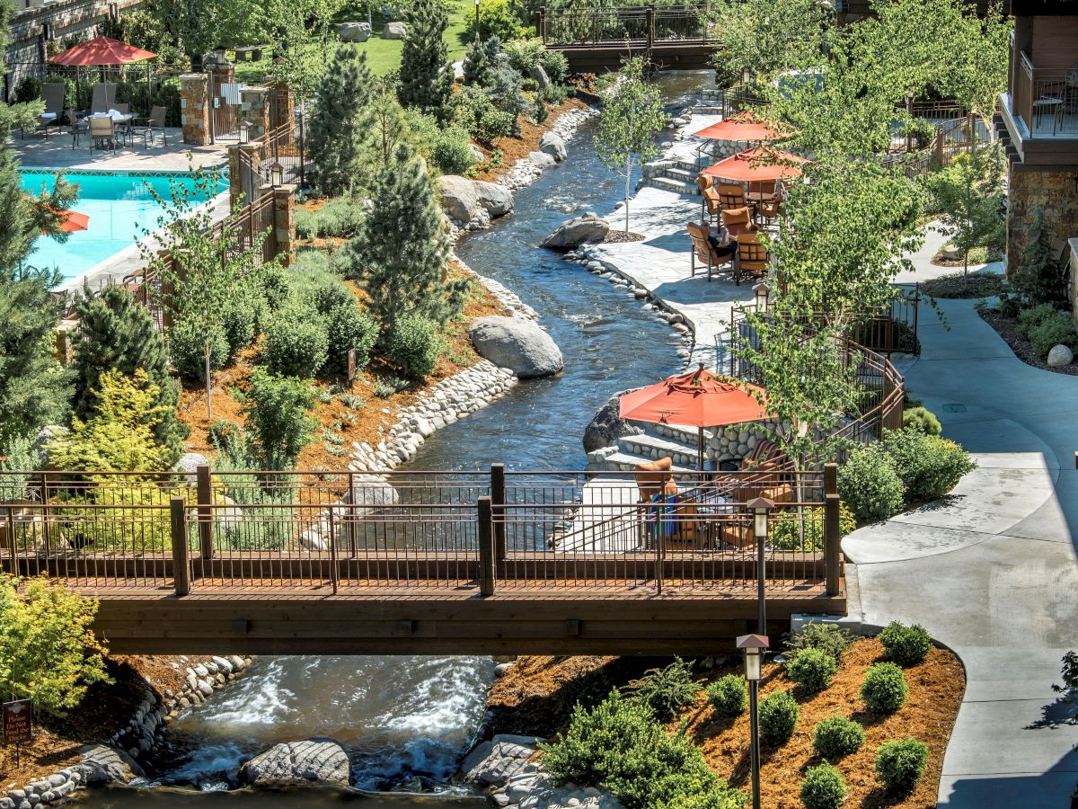 The image shows a resort area with a stream, footbridges, greenery, a swimming pool, and outdoor seating with red umbrellas, creating a serene atmosphere.