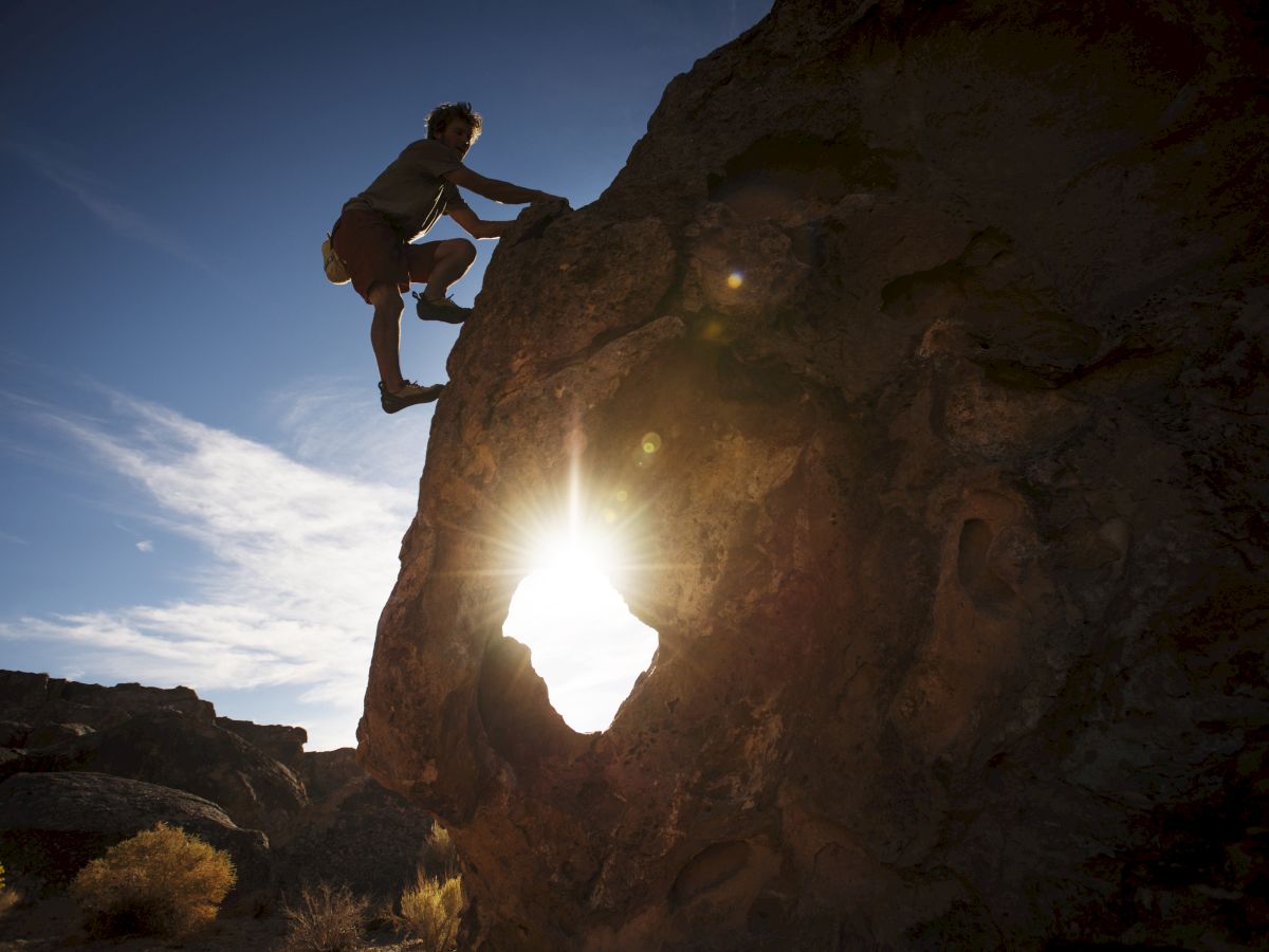 A person is rock climbing in a mountainous area with the sun shining through a hole in the rock face. The sky is clear with some clouds.