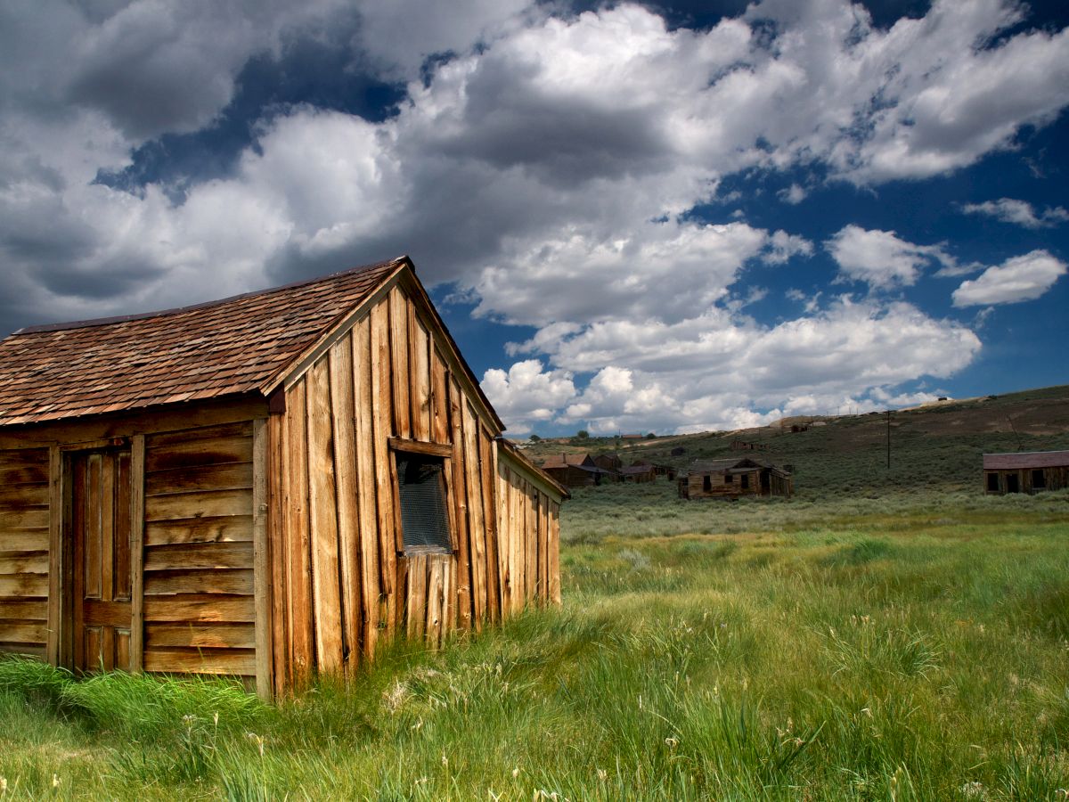 An old wooden cabin stands in a grassy field under a cloudy sky, with additional structures visible in the distant background.