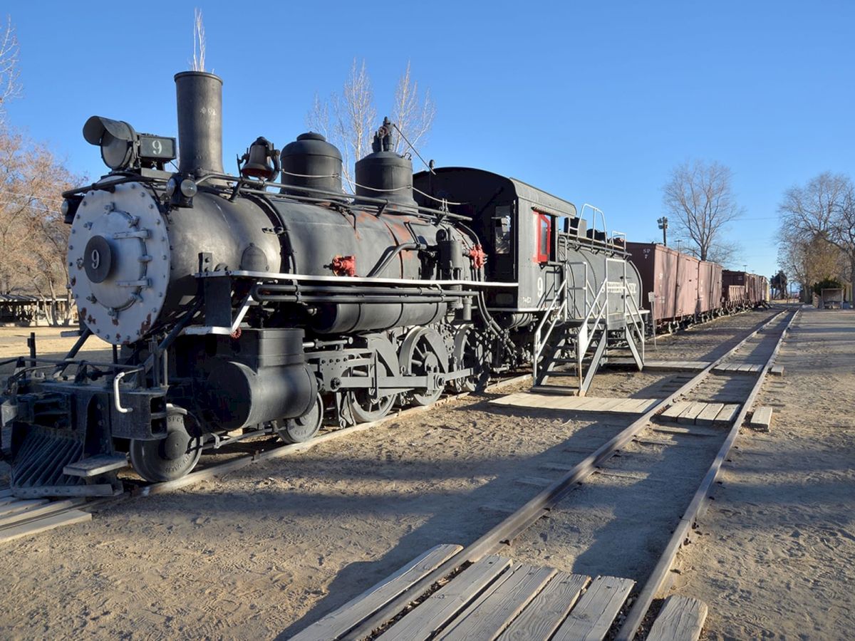 The image shows an old-fashioned steam locomotive on railway tracks, with some cargo cars in the background and a clear blue sky.