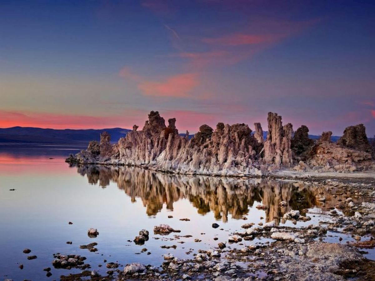 A sunset view of a serene lake with unique, rocky formations reflecting on the still water, with a mix of pink and blue hues in the sky ending the sentence.