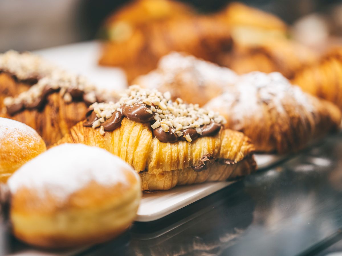 The image shows an assortment of pastries, including croissants topped with chocolate and nuts, and other pastries dusted with powdered sugar.