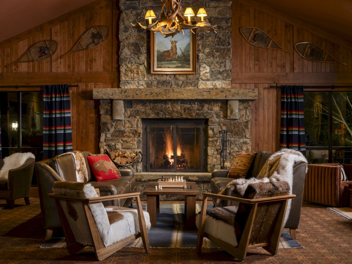 A cozy rustic living room features a stone fireplace, wooden walls, and comfortable seating arranged around a chessboard, with warm lighting.