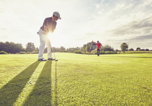 Two people are playing golf on a sunny day. One is putting the ball while the other stands near the flag on the green.