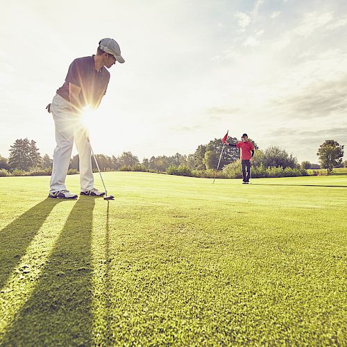 Two people are playing golf on a sunny day. One is putting the ball while the other stands near the flag on the green.