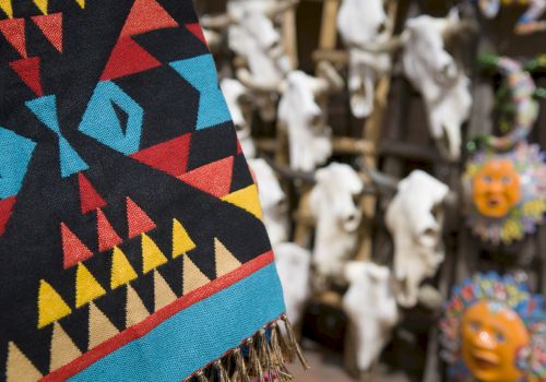 The image shows a colorful, patterned textile in the foreground, with animal skulls and decorative masks in the blurry background.